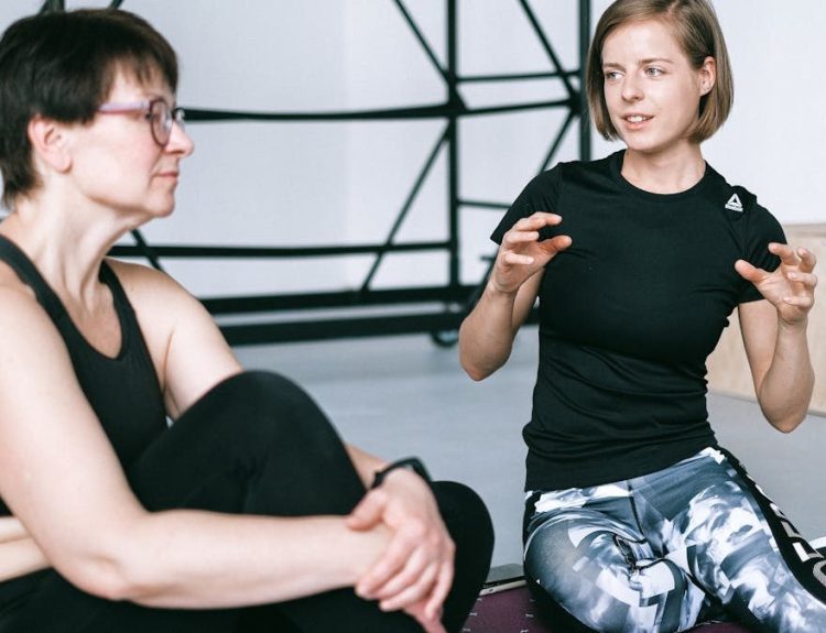Two women in sportswear sitting on a yoga mat, having a detailed discussion in a gym.