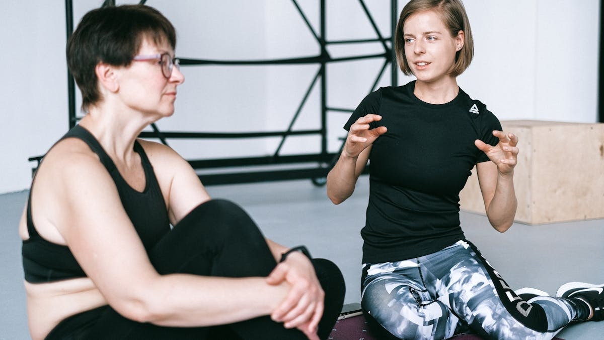 Two women in sportswear sitting on a yoga mat, having a detailed discussion in a gym.